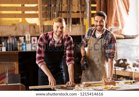 profession, carpentry, woodwork and people concept - two happy carpenters with electric drill and wooden board at workshop