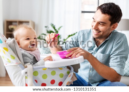 family, food, eating and people concept - happy father feeding little baby daughter sitting in highchair with puree by spoon at home