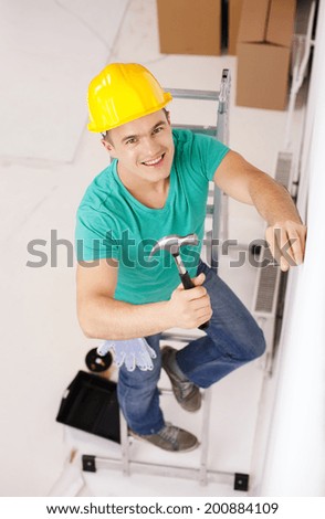 repair, building and home renovation concept - smiling man in yellow protective helmet hammering nail in wall