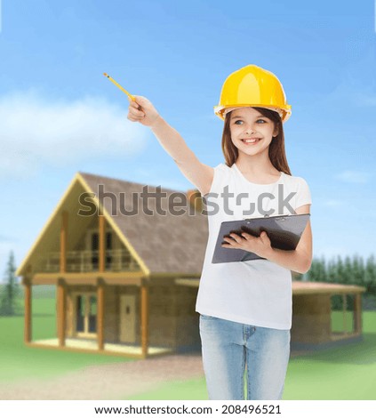 childhood, construction, architecture, building and people concept - smiling little girl in protective helmet with clipboard pointing pencil