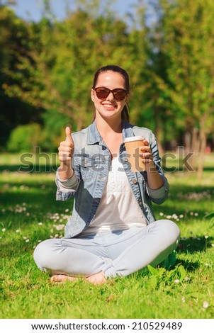 lifestyle, summer vacation, drinks and people concept - smiling young girl drinking coffee from paper cup and showing thumbs up in park