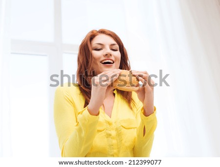 health, eating, food, home and people concept - smiling young woman eating hamburger at home