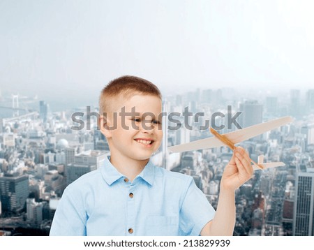 dreams, future, hobby, urban life and childhood concept - smiling little boy holding wooden airplane model in his hand over city background