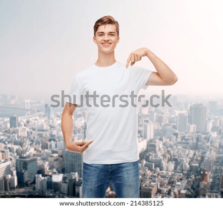 gesture, advertising and people concept - smiling young man in blank white t-shirt pointing fingers on himself over city background