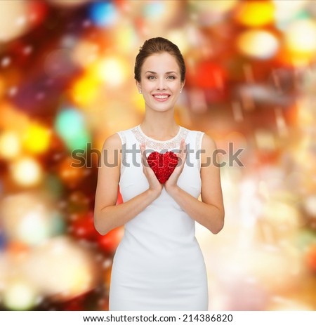 happiness, health, charity and love concept - smiling woman in white dress with red heart over party lights background