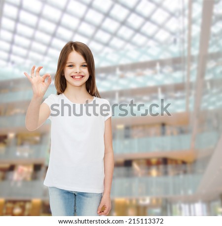 advertising, childhood, gesture, consumerism and people - smiling little girl in white blank t-shirt showing ok sign over shopping center background