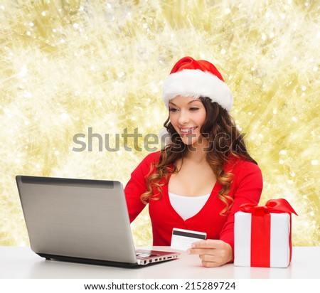 christmas, holidays, technology and shopping concept - smiling woman in santa helper hat with gift box, credit card and laptop computer over yellow lights background