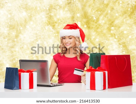 christmas, holidays, technology and shopping concept - smiling woman in santa helper hat with gifts, credit card and laptop computer over yellow lights background