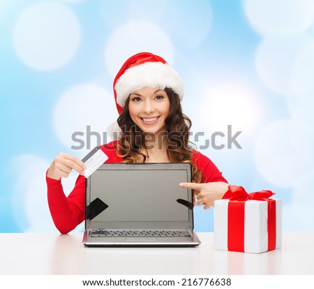 christmas, holidays, technology and shopping concept - smiling woman in santa helper hat with gift box, credit card and laptop computer pointing finger over blue lights background