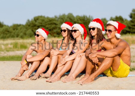 friendship, happiness, summer vacation, holidays and people concept - group of friends in santa helper hats sitting on beach