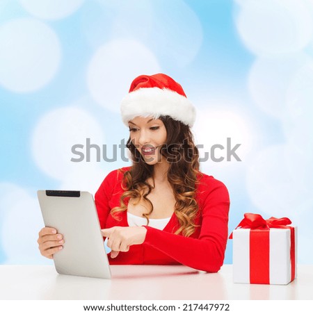 christmas, holidays, technology and people concept - smiling woman in santa helper hat with gift box and tablet pc computer over blue lights background