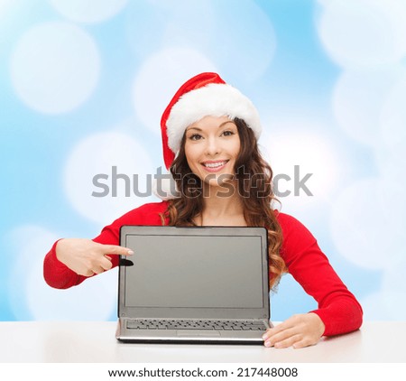 christmas, holidays, technology, advertisement and people concept - smiling woman in santa helper hat pointig finger to blank laptop computer screen over blue lights background