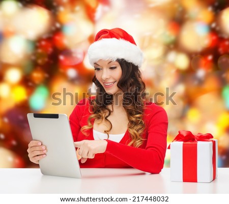 christmas, holidays, technology and people concept - smiling woman in santa helper hat with gift box and tablet pc computer over red lights background