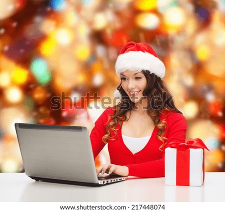 christmas, holidays, technology and people concept - smiling woman in santa helper hat with gift box and laptop computer over red lights background