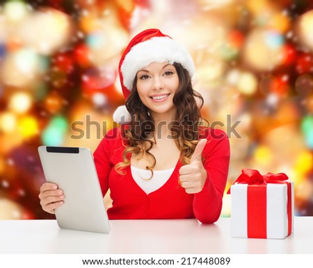 christmas, holidays, technology, gesture and people concept - smiling woman in santa helper hat with gift box and tablet pc computer showing thumbs up over red lights background