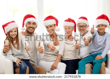 family, happiness, generation, holidays and people concept - happy family in santa helper hats sitting on couch and showing thumbs up gesture at home