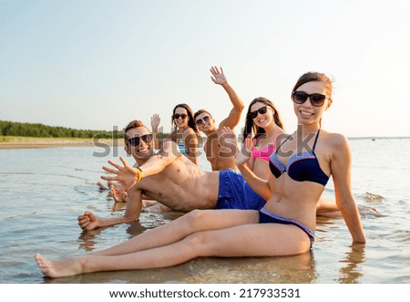 friendship, sea, holidays, gesture and people concept - group of smiling friends wearing swimwear and sunglasses sitting in water on beach