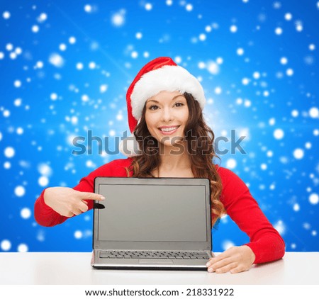 christmas, holidays, technology, advertisement and people concept - smiling woman in santa helper hat pointig finger to blank laptop computer screen over blue snowing background