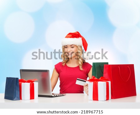 christmas, holidays, technology and shopping concept - smiling woman in santa helper hat with gifts, credit card and laptop computer over blue lights background