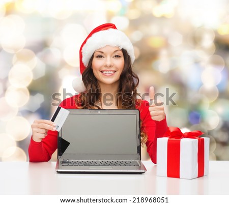 christmas, holidays, technology and shopping concept - smiling woman in santa helper hat with credit card, gift box and laptop computer showing thumbs up gesture over lights background
