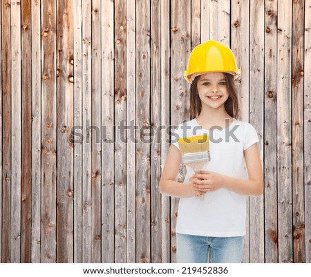painting, building, childhood and people concept - smiling little girl in protective helmet with paint brush over wooden fence background