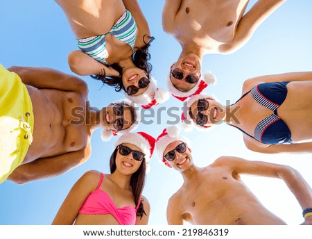 friendship, christmas, summer vacation, holidays and people concept - group of smiling friends wearing swimwear and santa helper hats standing in circle over blue sky