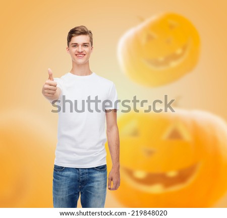 holidays, advertisement, gesture and people concept - smiling young man in blank white t-shirt showing thumbs up over halloween pumpkins background