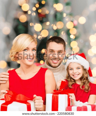 christmas, holidays,  family and people concept - happy mother, father and little girl in santa helper hat with gift boxes over over living room and christmas tree background