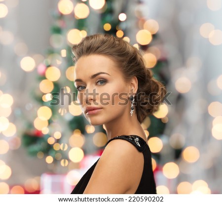 people, holidays, christmas and glamour concept - beautiful woman in evening dress wearing earrings over christmas tree and lights background