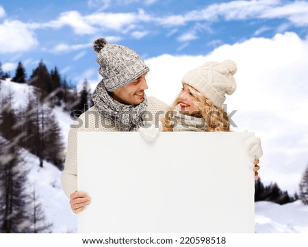 winter, holidays, christmas, advertisement and people concept - smiling couple in winter clothes with white blank billboard over snowy mountains background