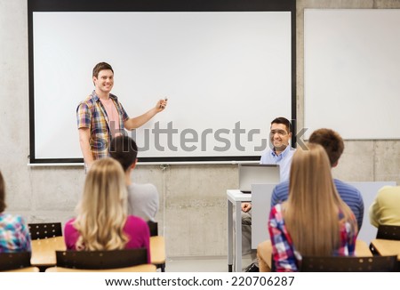 education, high school, technology and people concept - smiling student boy with remote control, laptop computer standing in front of white board and teacher in classroom