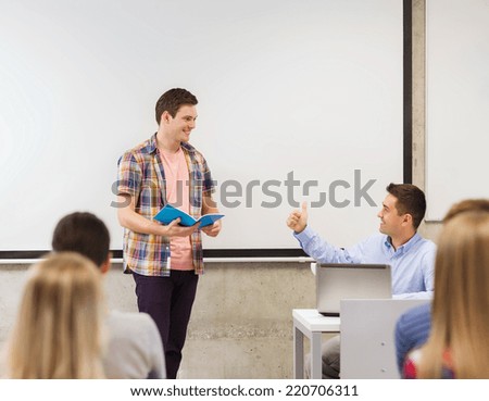 education, high school, technology and people concept - smiling student boy with notebook, laptop computer standing in front of students and teacher showing thumbs up gesture in classroom