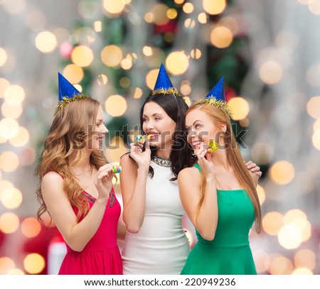 holidays, people and celebration concept - smiling women in party caps blowing to whistles over christmas tree lights background