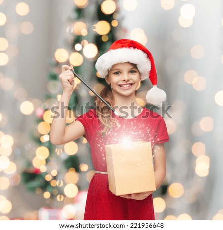 christmas, holidays, happiness and people concept - smiling girl in santa helper hat with gift box and magic wand over living room and christmas tree background