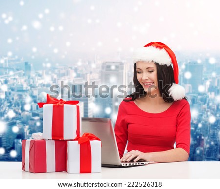 christmas, holidays, technology and people concept - smiling woman in santa helper hat with gift boxes and laptop computer over snowy city background