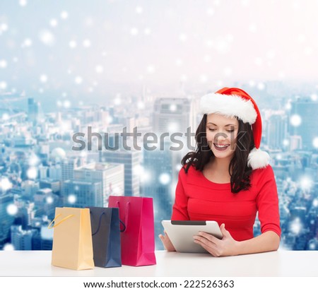 christmas, holidays, technology and people concept - smiling woman in santa helper hat with shopping bags and tablet pc computer over snowy city background