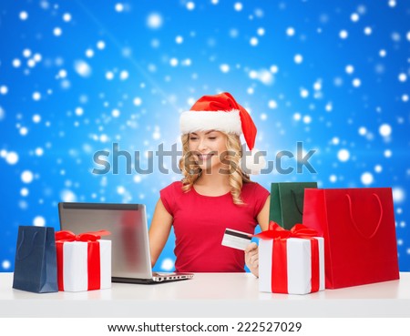 christmas, holidays, technology and shopping concept - smiling woman in santa helper hat with gifts, credit card and laptop computer over blue snowy background