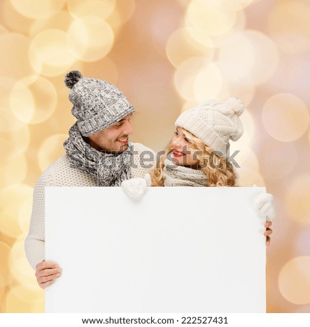 winter, holidays, christmas, advertisement and people concept - smiling couple in winter clothes with white blank billboard over beige lights background