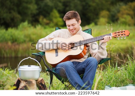 adventure, travel, tourism and people concept - smiling man with guitar and cooking food on bonfire in camping