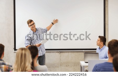 education, high school, technology and people concept - student standing with remote control, laptop computer in front of teacher and classmates in classroom