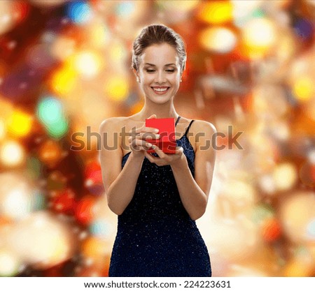 winter holidays, christmas, presents, luxury and people concept - smiling woman in dress holding red gift box over red lights background