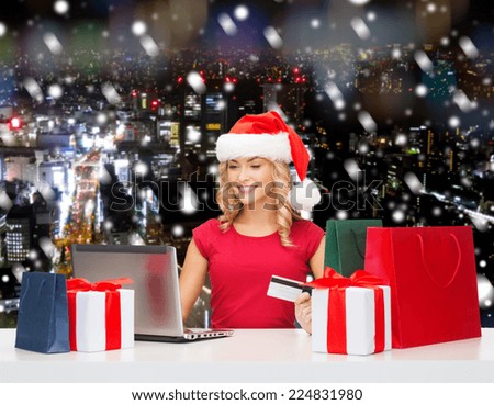 christmas, holidays, technology and shopping concept - smiling woman in santa helper hat with gifts, credit card and laptop computer over blue snowy background