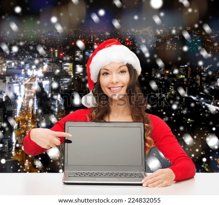 christmas, holidays, technology, advertisement and people concept - smiling woman in santa helper hat pointig finger to blank laptop computer screen over snowy night city background