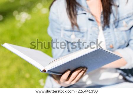 lifestyle, summer vacation, education, literature and people concept - close up of young girl reading book and sitting on grass in park