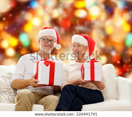 family, holidays, christmas, age and people concept - happy senior couple in santa helper hats with gift boxes over red lights background