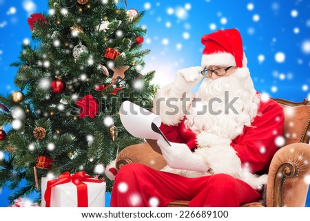 christmas, holidays and people concept - man in costume of santa claus with notepad and christmas tree sitting in armchair over blue snowy background