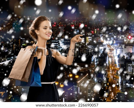 shopping, sale, christmas, people and holidays concept - smiling woman in evening dress with shopping bags pointing finger over snowy night city background