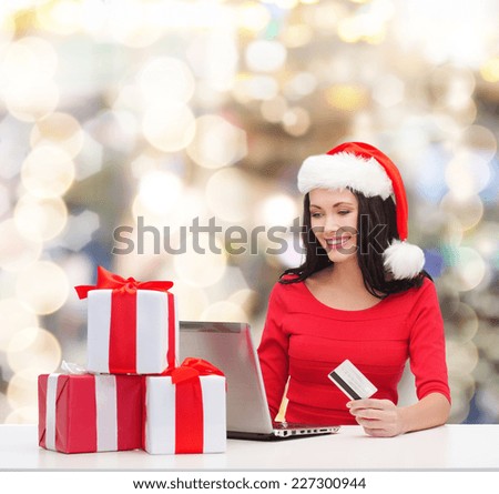 christmas, shopping and people concept - smiling woman in santa helper hat with gift boxes, laptop computer and credit card over lights background