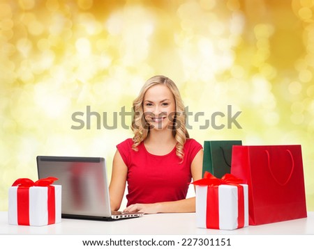 christmas, holidays, technology, advertising and people concept - smiling woman in red blank shirt with shopping bags, gifts and laptop computer over yellow lights background