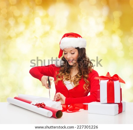 christmas, holidays, celebration, decoration and people concept - smiling woman in santa helper hat with scissors packing gift box over yellow lights background